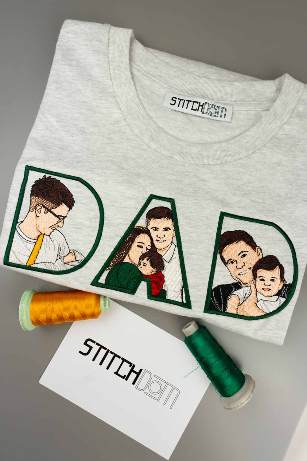 Tricou gri deschis din bumbac organic, împăturit, cu textul „DAD” brodat personalizat, cu poze de familie brodate în interiorul literelor, pe o suprafață gri, alături de conuri de ață și un card „Stitchdom”.