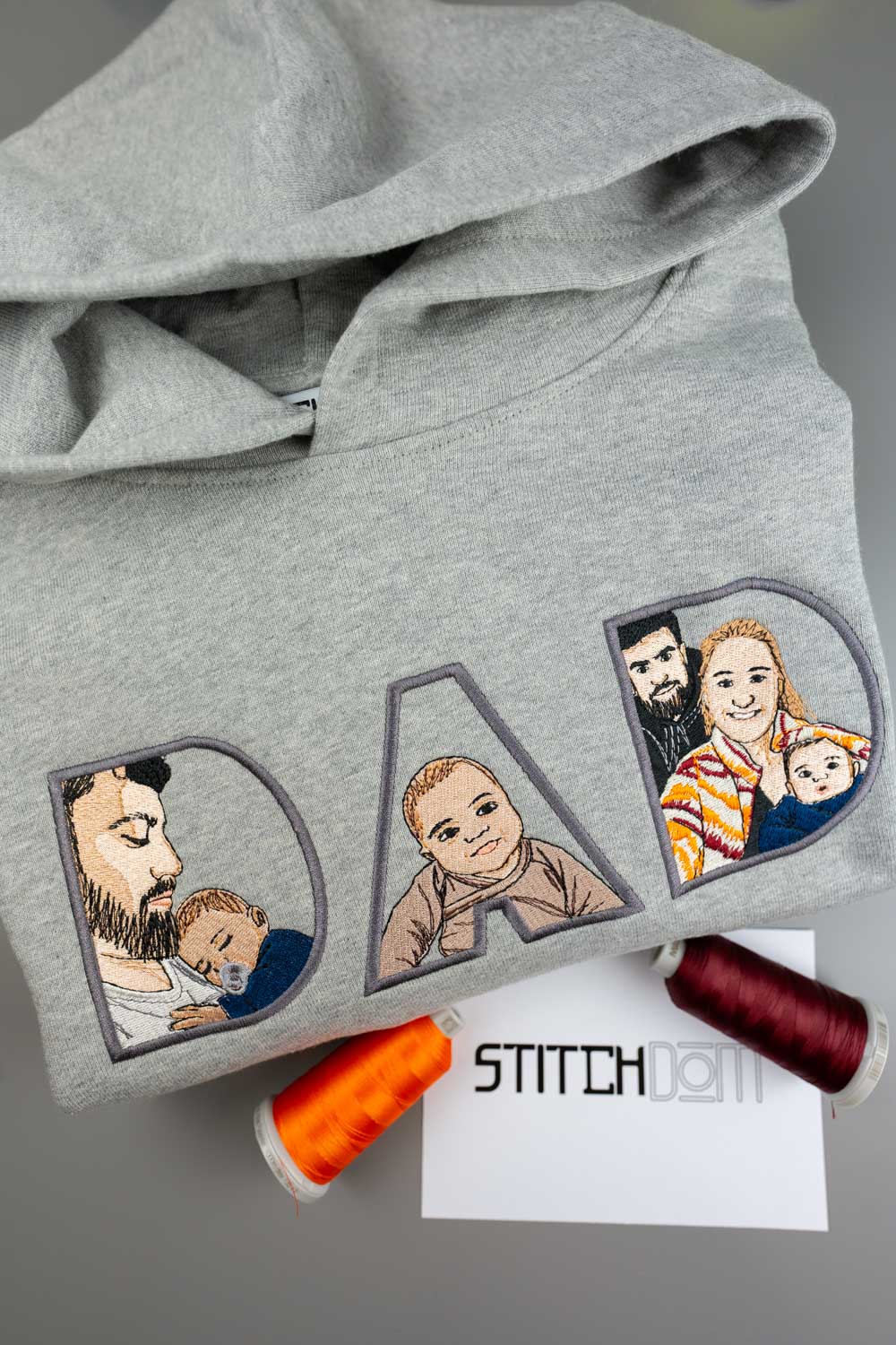 Hanorac gri deschis din bumbac organic, așezat flat lay pe o suprafață gri, cu literele „DAD” brodate și portrete ilustrate brodate în interiorul fiecărei litere, alături de conuri de ață portocalie și bordo și un mic card cu textul „STITCHDOM”.