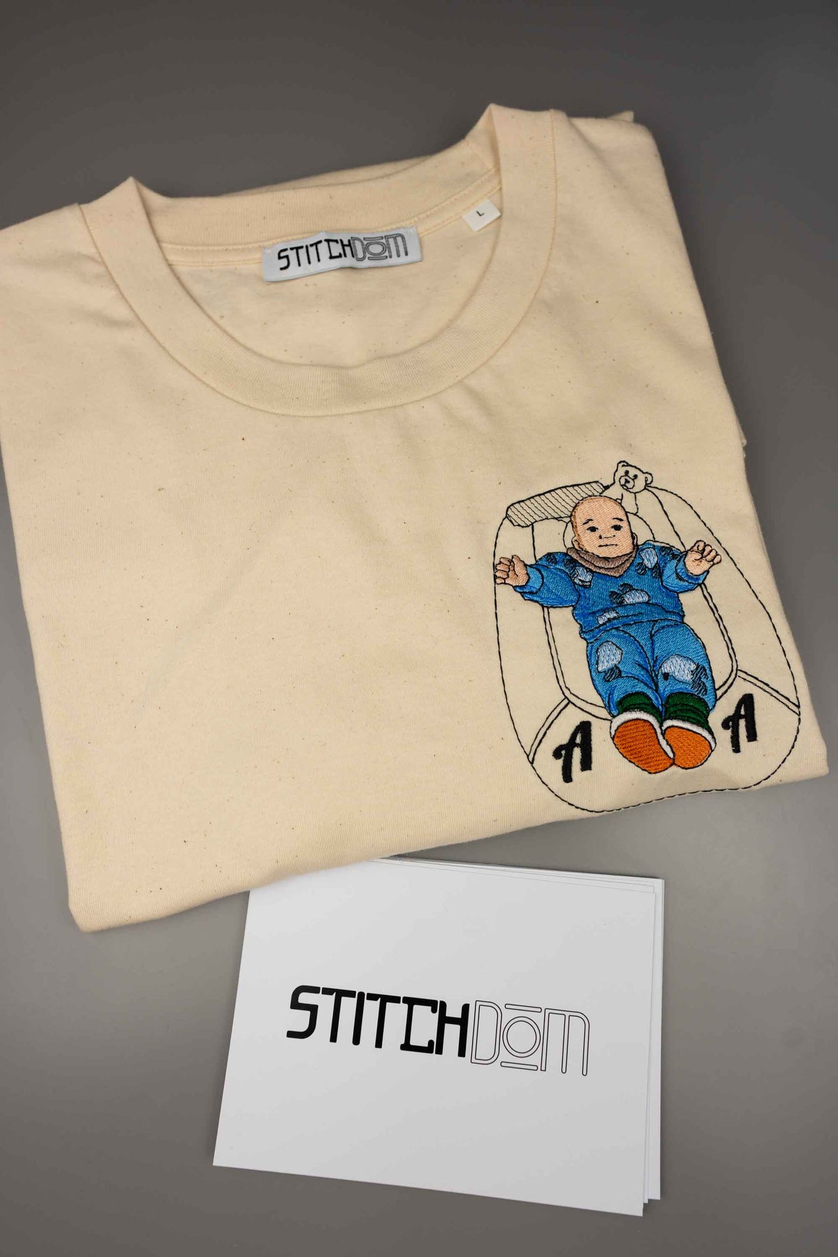 Tricou bej din bumbac organic împăturit, cu un model brodat reprezentând un bebeluș care stă în pătuțul lui, cu brandul „Stitchdom” vizibil, pe o suprafață gri.