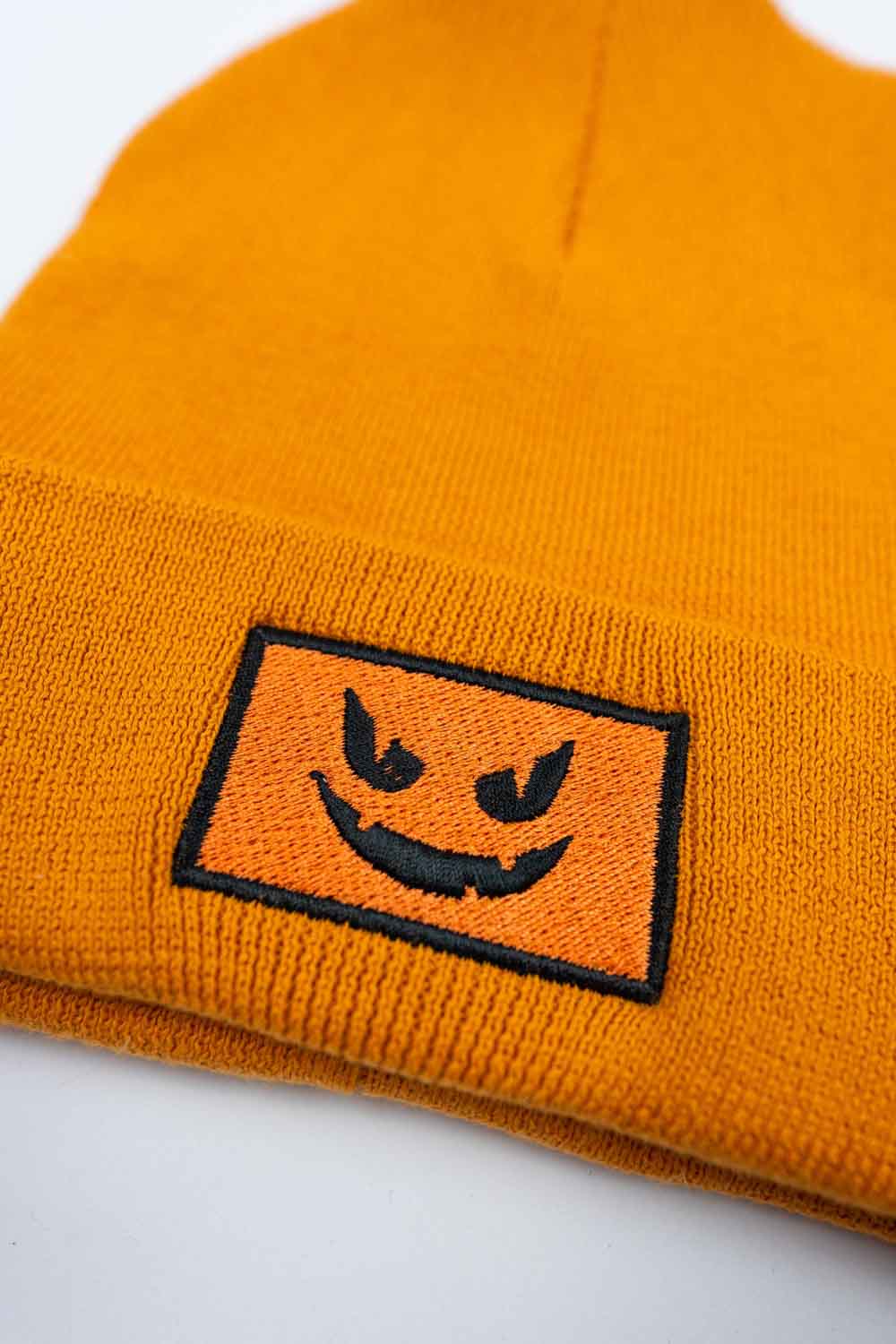 Căciulă portocalie din tricot reiat cu manșetă întoarsă, brodată cu o față de dovleac Jack-o'-lantern neagră într-un chenar dreptunghiular, un accesoriu amuzant inspirat de Halloween.