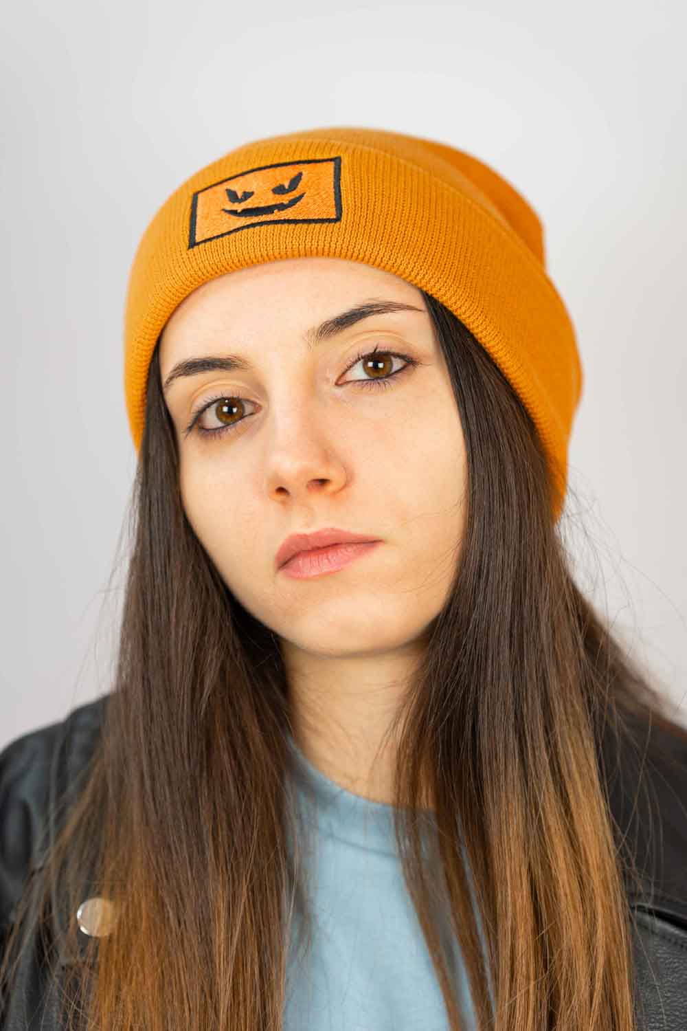 Căciulă portocalie din tricot reiat cu manșetă întoarsă, brodată cu o față de dovleac Jack-o'-lantern neagră într-un chenar dreptunghiular, un accesoriu amuzant inspirat de Halloween.