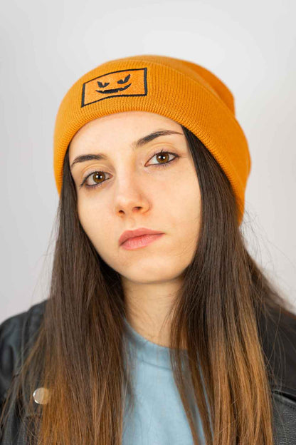 Căciulă portocalie din tricot reiat cu manșetă întoarsă, brodată cu o față de dovleac Jack-o'-lantern neagră într-un chenar dreptunghiular, un accesoriu amuzant inspirat de Halloween.