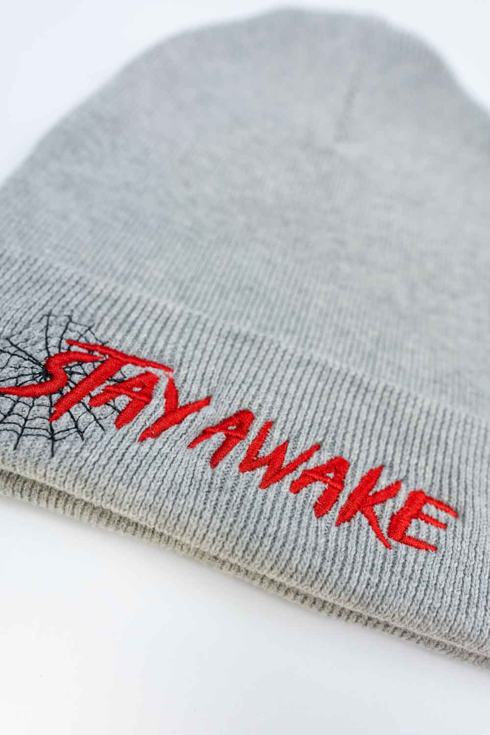Căciulă gri din tricot cu manșetă întoarsă, brodată cu mesajul „Stay Awake” în roșu și un mic design de pânză de păianjen neagră, un accesoriu original și amuzant pentru un stil casual.
