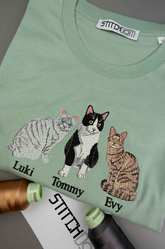 Tricou verde mentă cu mânecă scurtă din bumbac organic, așezat pe o suprafață gri, cu poze brodate cu trei pisici și numele „Luki”, „Tommy” și „Evy”, alături de conuri de ață și o etichetă albă.