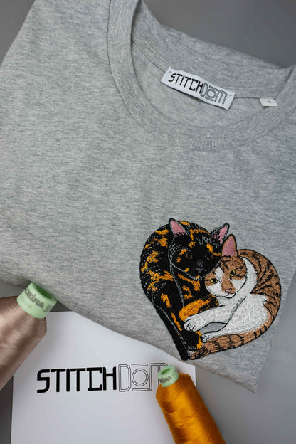 Tricou gri din bumbac organic brodat cu 2 pisici, una alba cu maro si una neagra cu portocaliu care se tin in brate in forma de inimia, pe un fundal gri cu un card cu brandingul "Stitchdom".