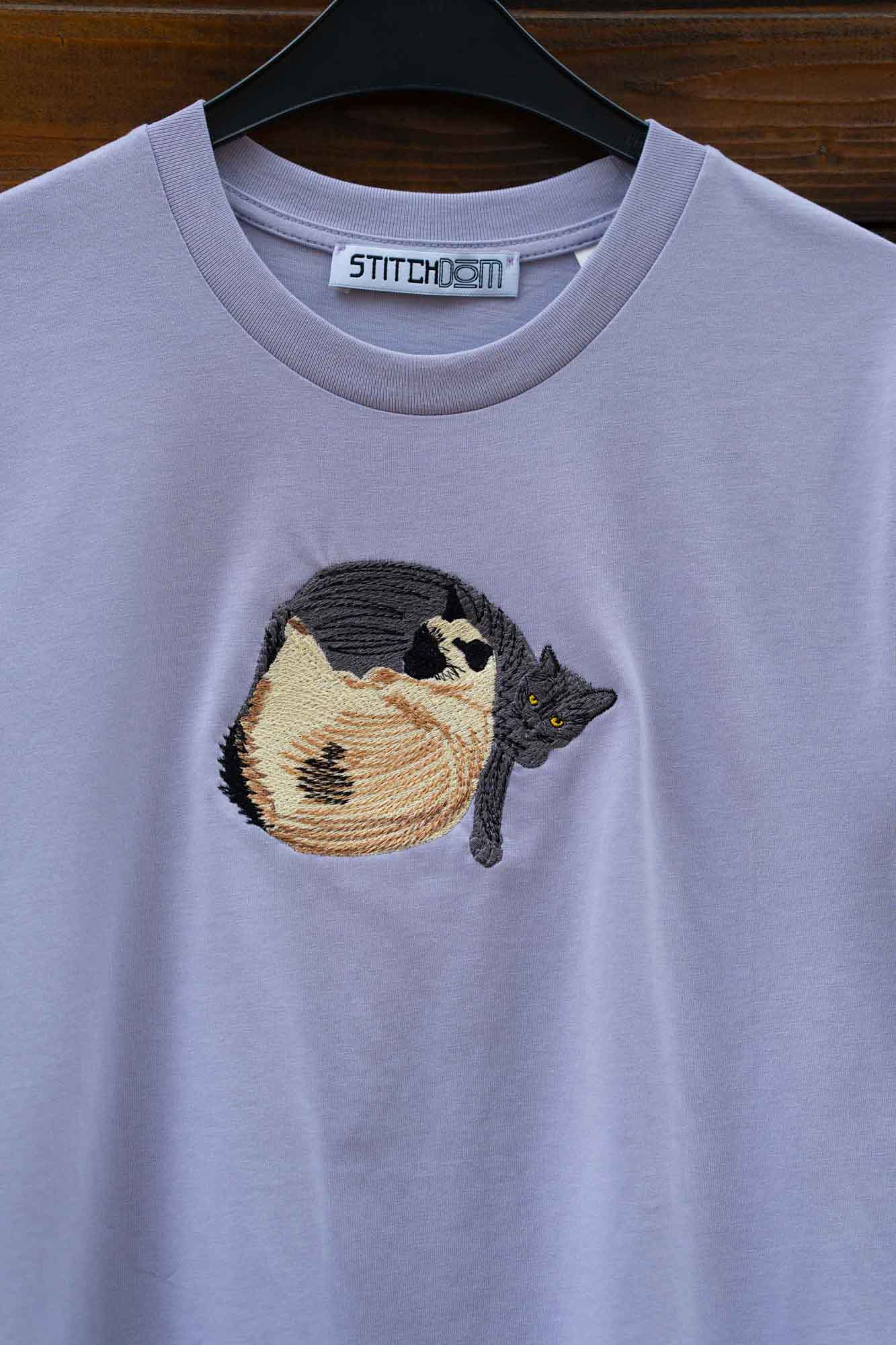 Tricou mov deschis cu model brodat reprezentând o pisică, pe umeraș.