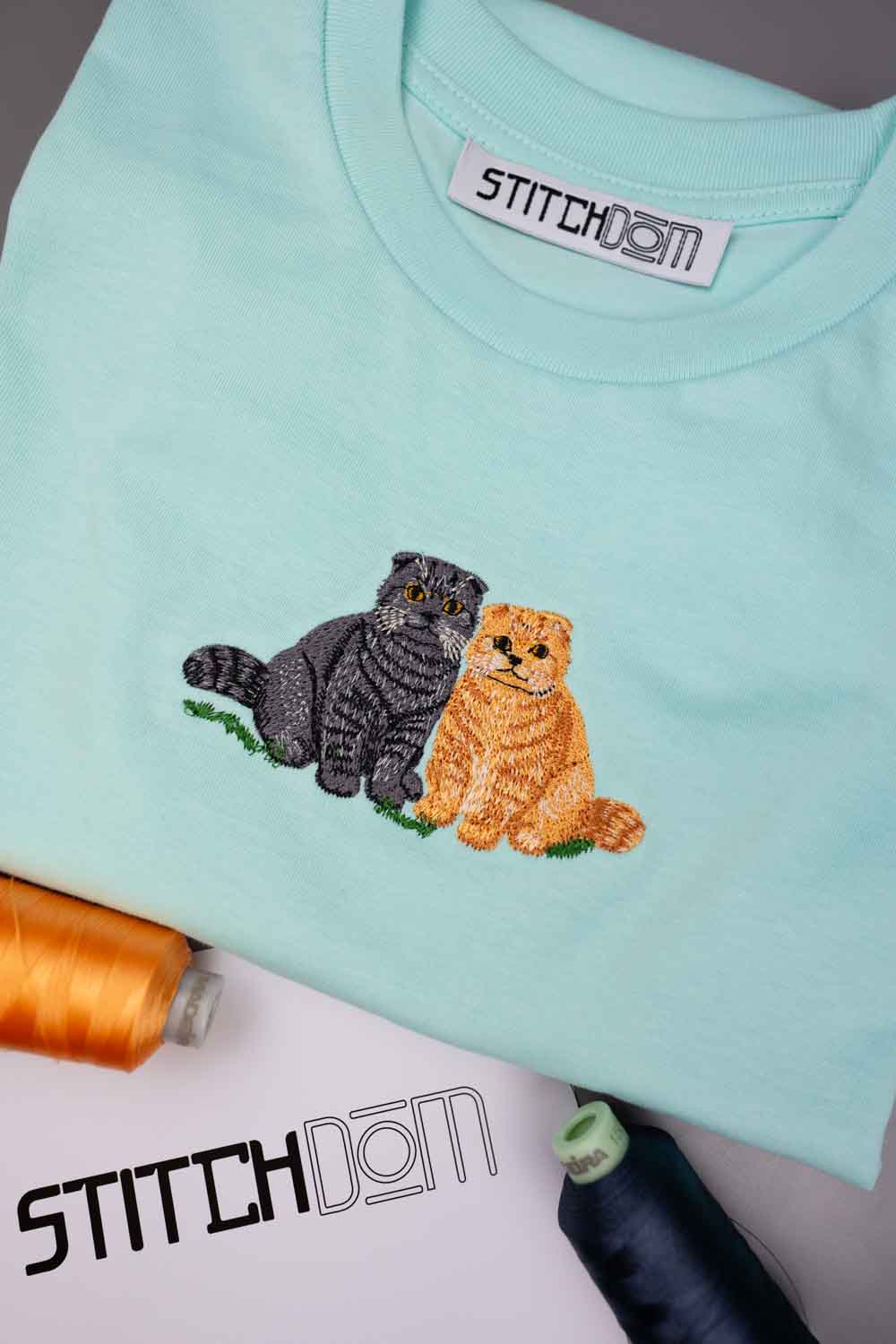Tricou verde mentă din bumbac organic, cu poze brodate cu o pisică tigrată neagră și o pisică tigrată portocalie așezate una lângă alta pe o bucată de iarbă verde, pe o suprafață albă, cu conuri de ață în partea din față și logo-ul „Stitchdom” vizibil pe eticheta gulerului și în partea stângă jos.