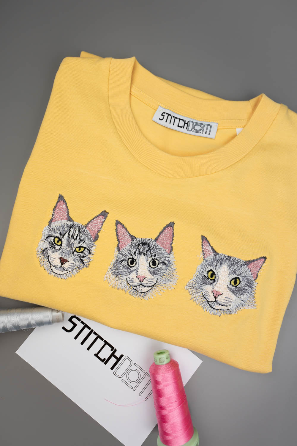 Tricou galben din bumbac organic, împăturit, cu trei poze brodate cu fețe de pisici pe piept, alături de accesorii de cusut, inclusiv un con de ață roz și o etichetă de brand, pe un fundal gri.