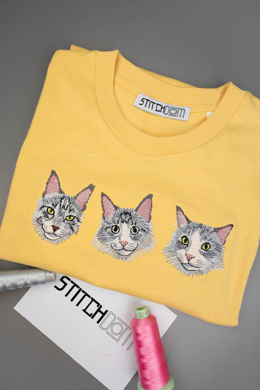Tricou galben din bumbac organic, împăturit, cu trei poze brodate cu fețe de pisici pe piept, alături de accesorii de cusut, inclusiv un con de ață roz și o etichetă de brand, pe un fundal gri.
