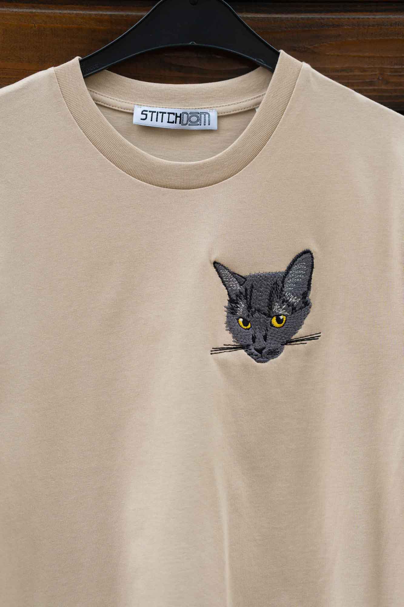 Tricou bej cu model brodat reprezentând fața unei pisici, pe umeraș.