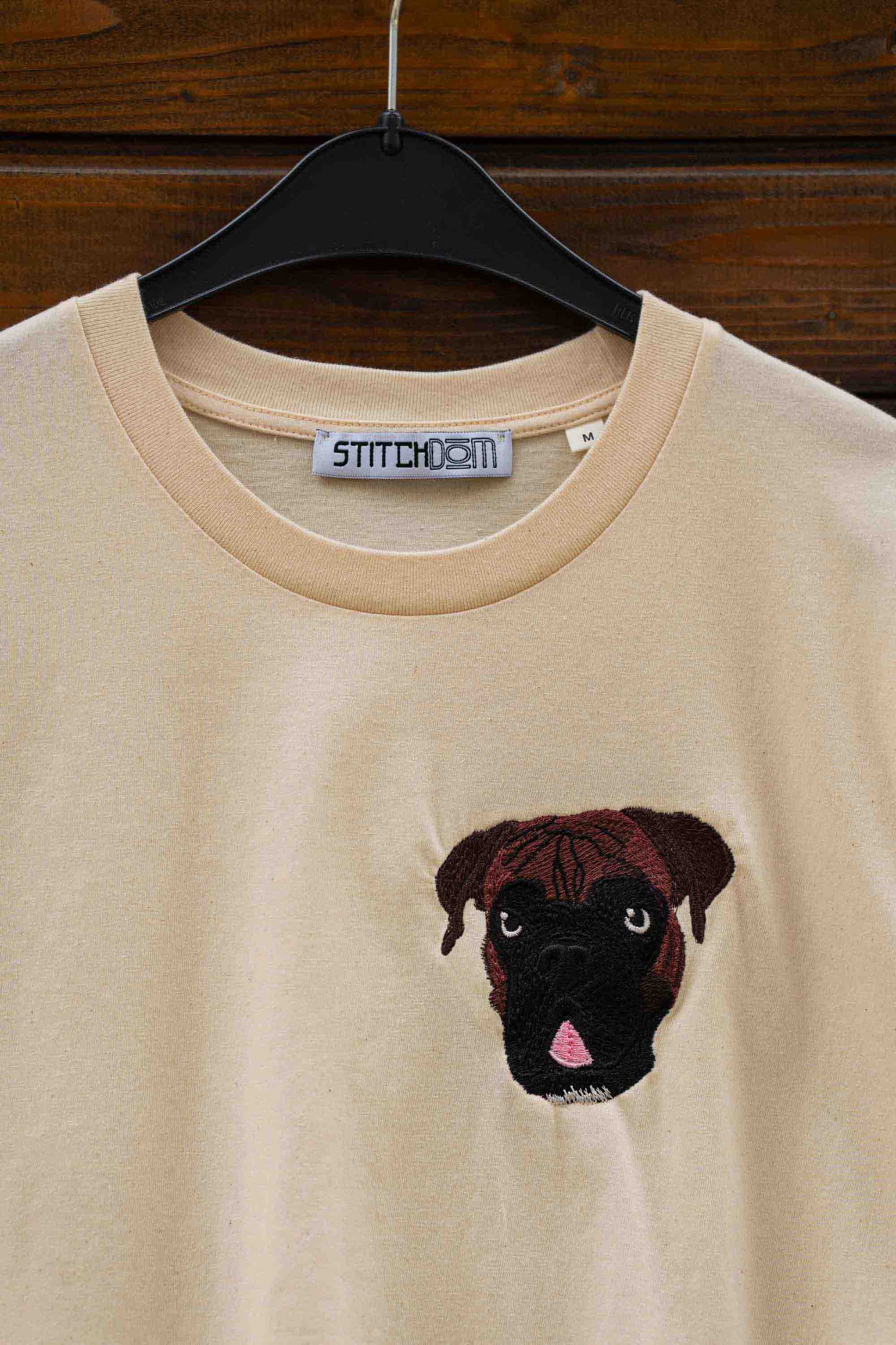 Tricou bej cu guler rotund, pe umeraș negru, pe fundal din lemn închis, cu un model brodat reprezentând fața unui câine Pug, pe partea stângă a pieptului.