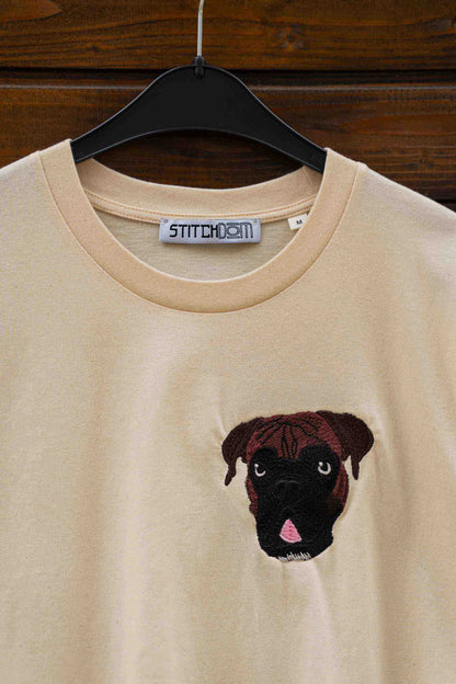 Tricou bej cu guler rotund, pe umeraș negru, pe fundal din lemn închis, cu un model brodat reprezentând fața unui câine Pug, pe partea stângă a pieptului.
