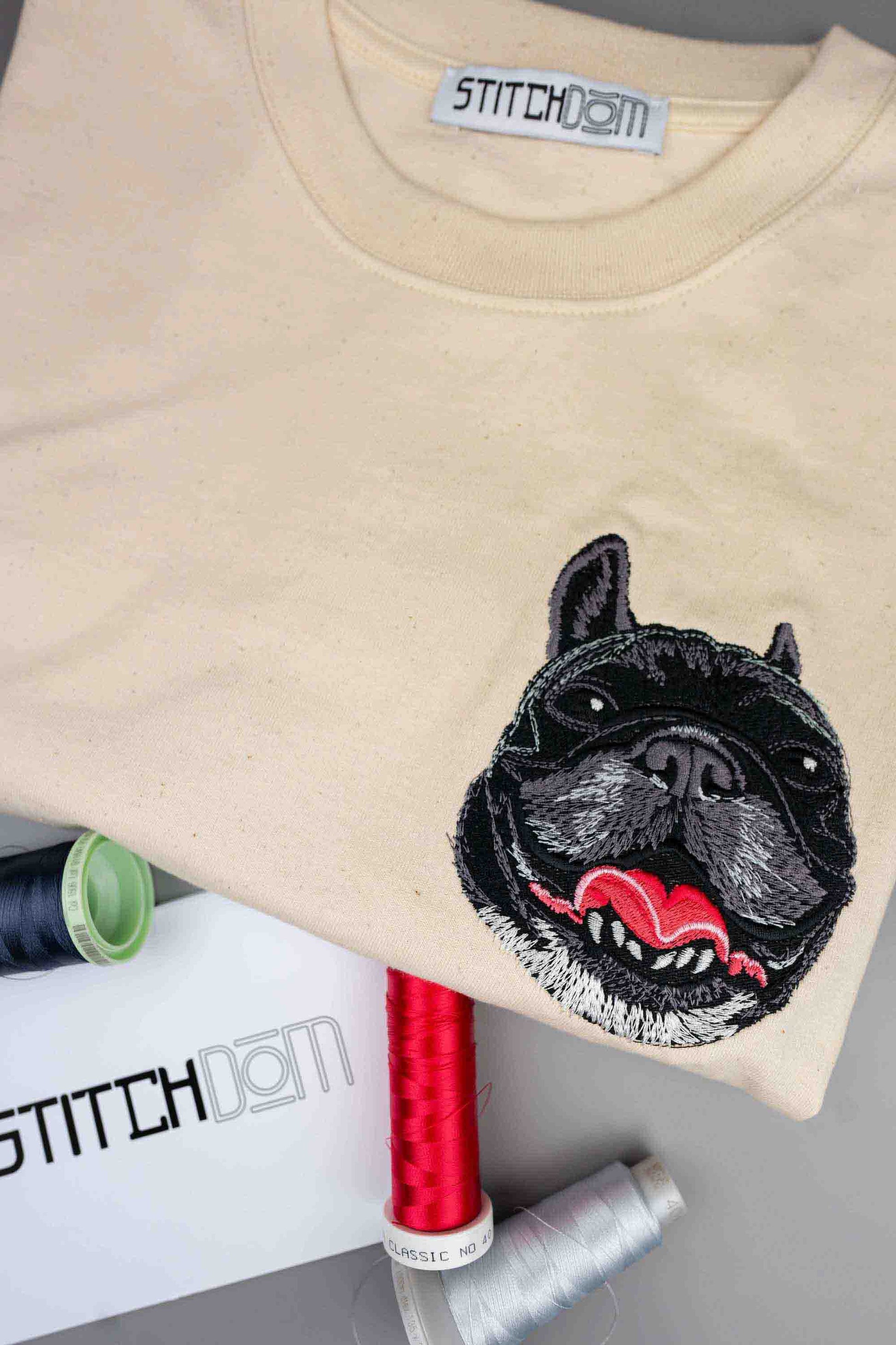 Tricou bej cu guler rotund, cu un model brodat reprezentând fața unui Bulldog Francez negru cu limba afara pe partea stângă a pieptului, așezat pe o suprafață gri, cu conuri de ață și un card branduit Stitchdom vizibile.