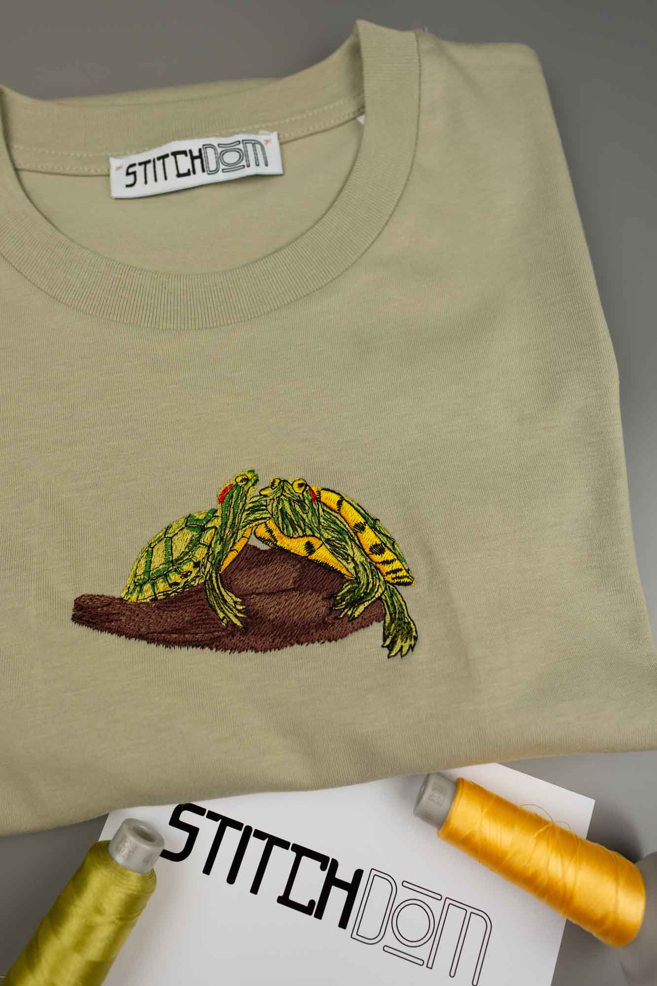 Tricou crem deschis din bumbac organic, împăturit, cu o poză brodată cu o broască verde și galbenă pe un buștean maro, cu eticheta „STITCHDOM” vizibilă în interiorul gulerului și conuri de ață în partea de jos.