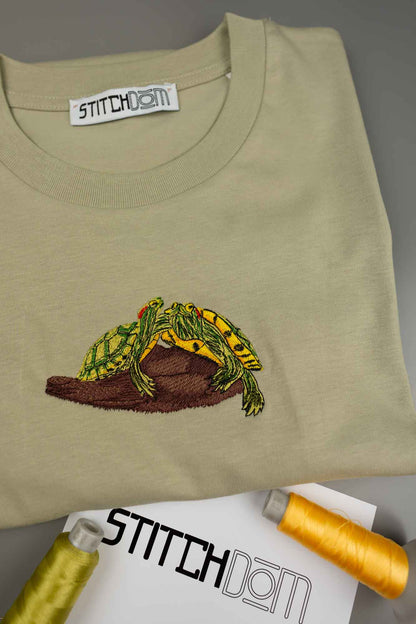 Tricou crem deschis din bumbac organic, împăturit, cu o poză brodată cu o broască verde și galbenă pe un buștean maro, cu eticheta „STITCHDOM” vizibilă în interiorul gulerului și conuri de ață în partea de jos.
