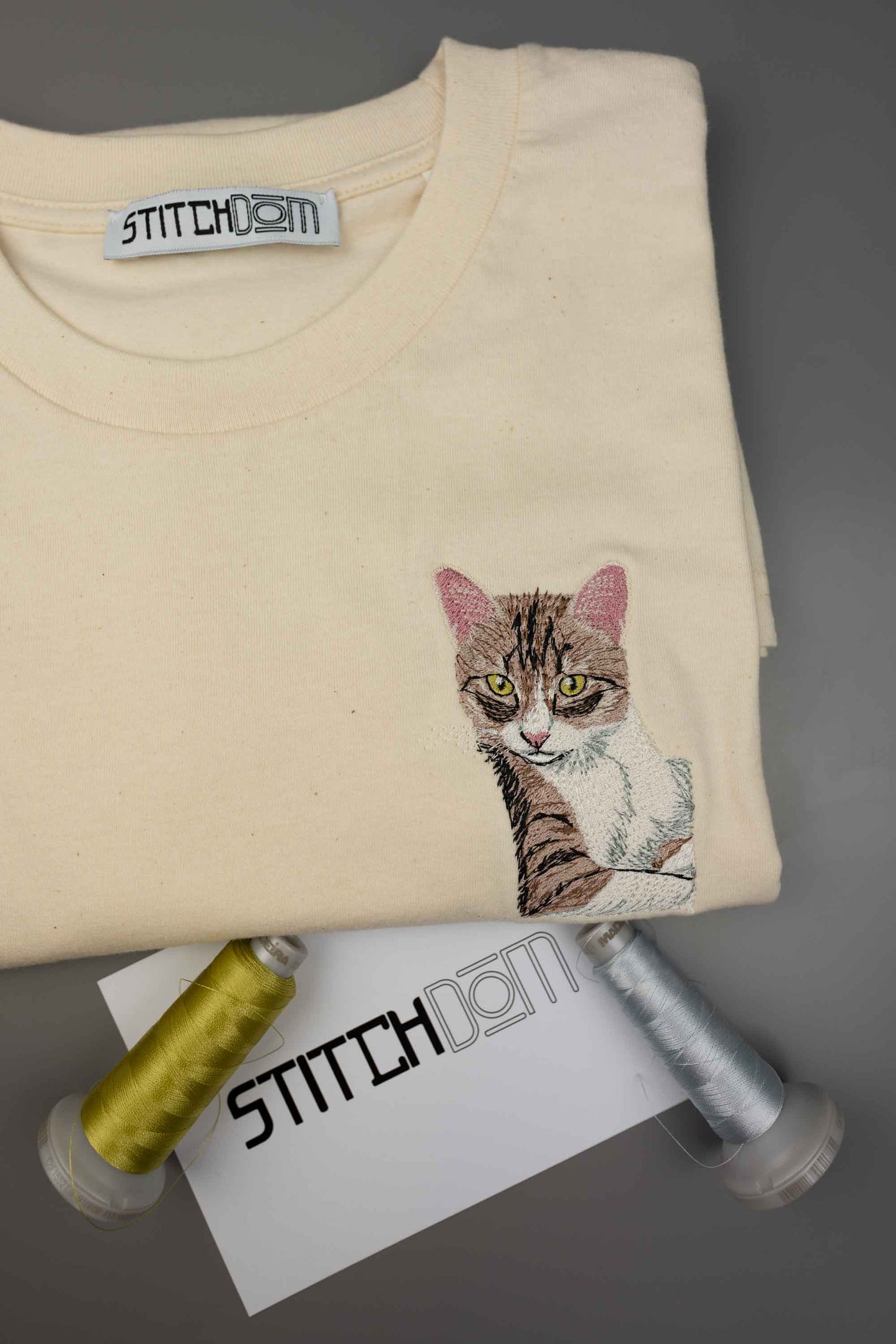 Tricou crem din bumbac organic, împăturit, de la Stitchdom, cu o poză brodată detaliată cu o pisică pe partea stângă a pieptului, alături de conuri de ață și un card de brand, pe un fundal gri.