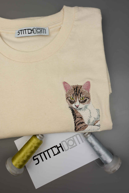 Tricou crem din bumbac organic, împăturit, de la Stitchdom, cu o poză brodată detaliată cu o pisică pe partea stângă a pieptului, alături de conuri de ață și un card de brand, pe un fundal gri.