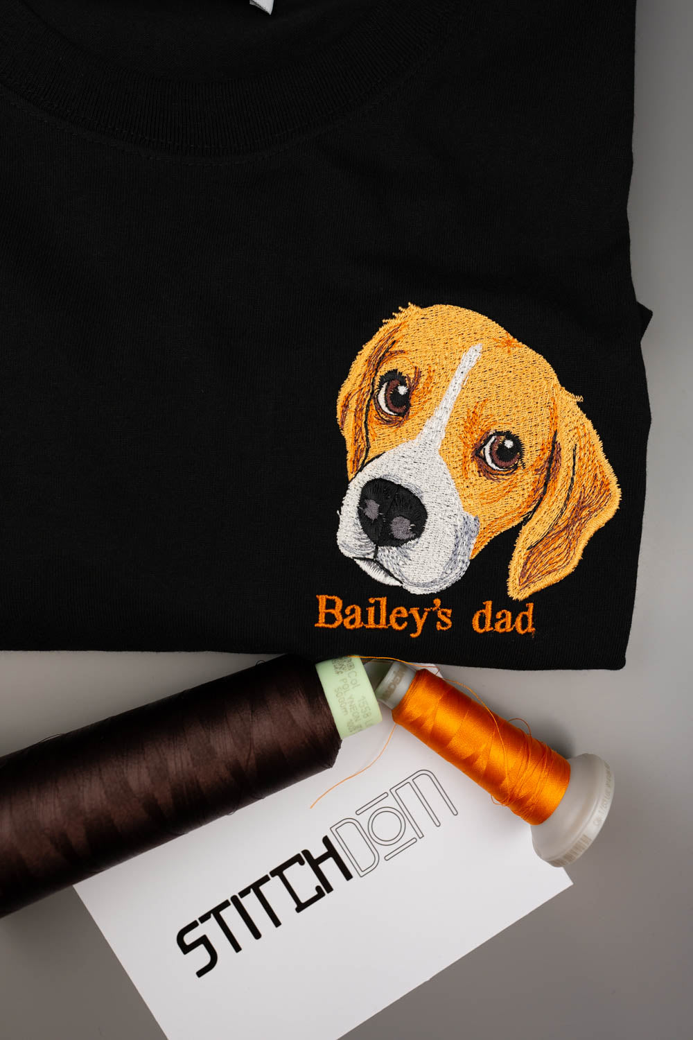 Tricou negru din bumbac organic, împăturit, cu o poză brodată cu fața unui câine beagle și textul „Bailey's dad” brodat cu portocaliu, alături de două conuri de ață și un card „Stitchdom”, pe o suprafață gri.