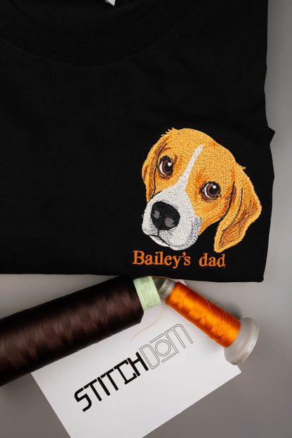 Tricou negru din bumbac organic, împăturit, cu o poză brodată cu fața unui câine beagle și textul „Bailey's dad” brodat cu portocaliu, alături de două conuri de ață și un card „Stitchdom”, pe o suprafață gri.