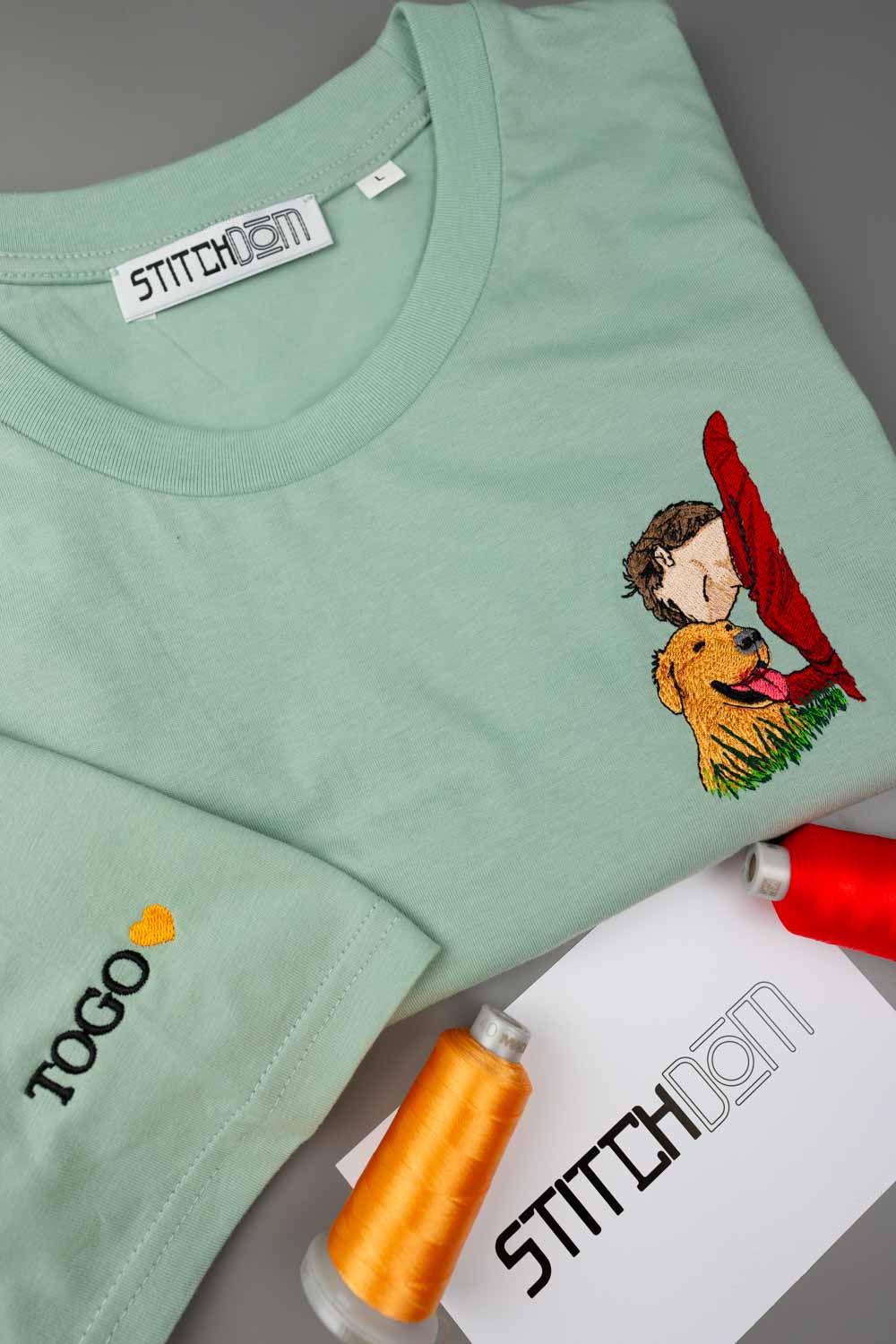 Tricou verde mentă cu mânecă scurtă din bumbac organic, așezat pe o suprafață gri, cu o poză brodată în stil desen cu o persoană care ține un steag roșu lângă un câine auriu, alături de conuri de ață și o etichetă de hârtie „Stitchdom”.