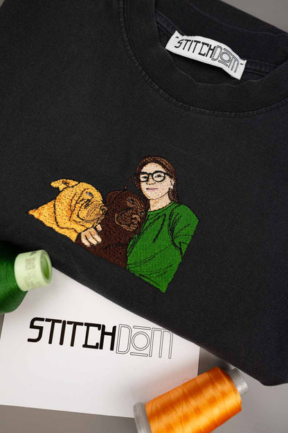 Tricou negru din bumbac organic, cu o poză brodată personalizată cu o femeie în pulover verde care îmbrățișează doi câini, unul auriu și unul maro, așezat pe o suprafață albă, cu eticheta „Stitchdom” vizibilă în interiorul gulerului și conuri de ață colorate alături.