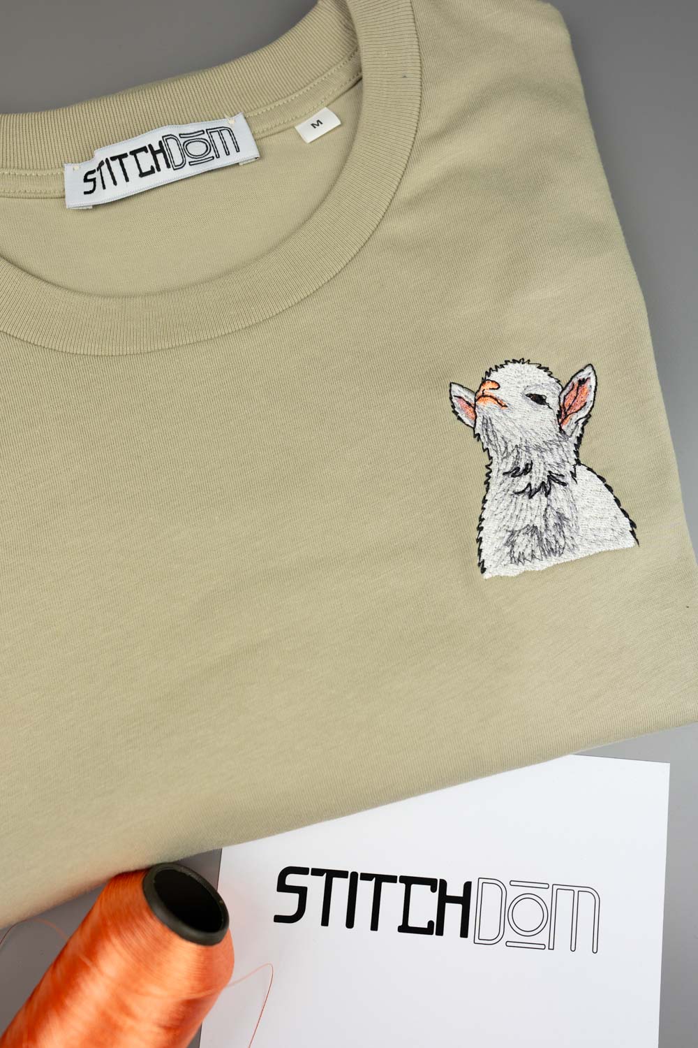 Tricou bej din bumbac organic, împăturit, cu o poză brodată alb-gri cu un miel pe partea stângă a pieptului, alături de un con de ață portocaliu și un card de brand „Stitchdom”, pe un fundal gri.