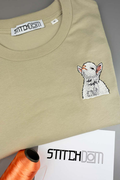 Tricou bej din bumbac organic, împăturit, cu o poză brodată alb-gri cu un miel pe partea stângă a pieptului, alături de un con de ață portocaliu și un card de brand „Stitchdom”, pe un fundal gri.