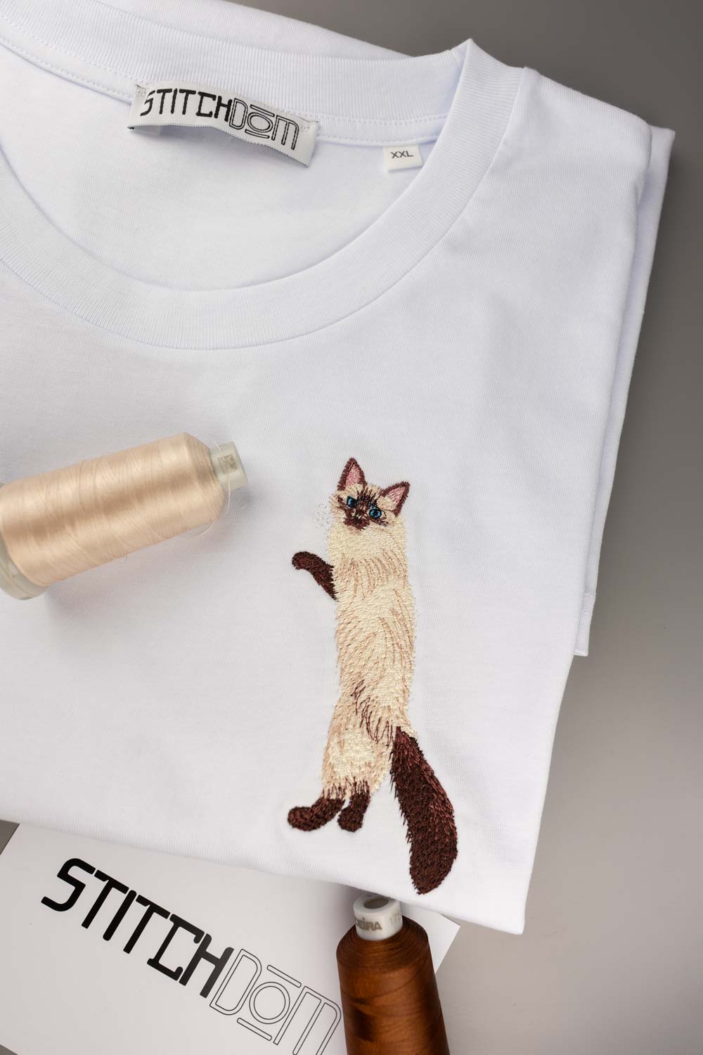 Tricou alb din bumbac organic, împăturit, cu o poză brodată detaliat cu o pisică siameză pe piept, alături de conuri de ață, pe un fundal gri neutru.