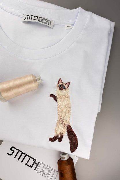 Tricou alb din bumbac organic, împăturit, cu o poză brodată detaliat cu o pisică siameză pe piept, alături de conuri de ață, pe un fundal gri neutru.