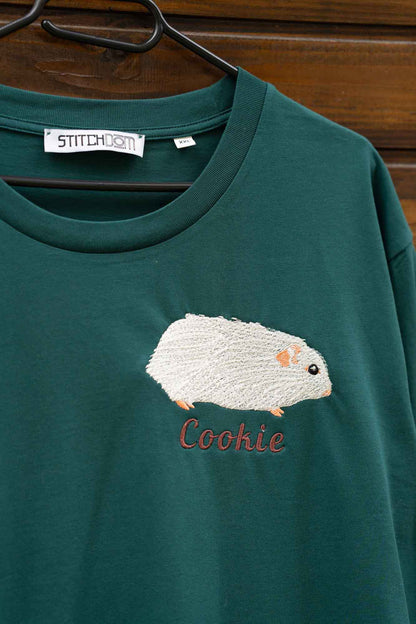 Tricou verde închis cu guler rotund, pe umeraș negru, pe fundal din lemn, cu un model brodat reprezentând un hamster alb și textul „Cookie” brodat cu scris roșu pe partea frontală.