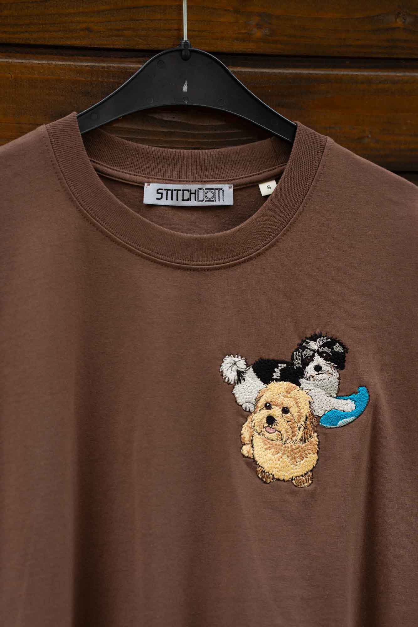 Tricou maro cu model brodat cu câini, pe umeraș, pe fundal din lemn.