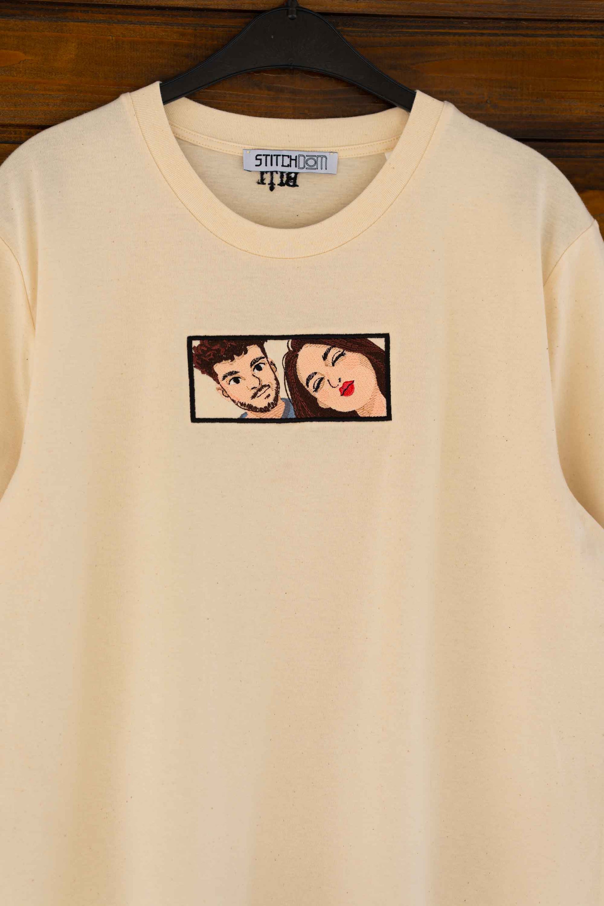 Tricou bej din bumbac organic, pe umeraș, pe fundal din lemn, cu un model brodat reprezentând un cuplu intr-un cadran.