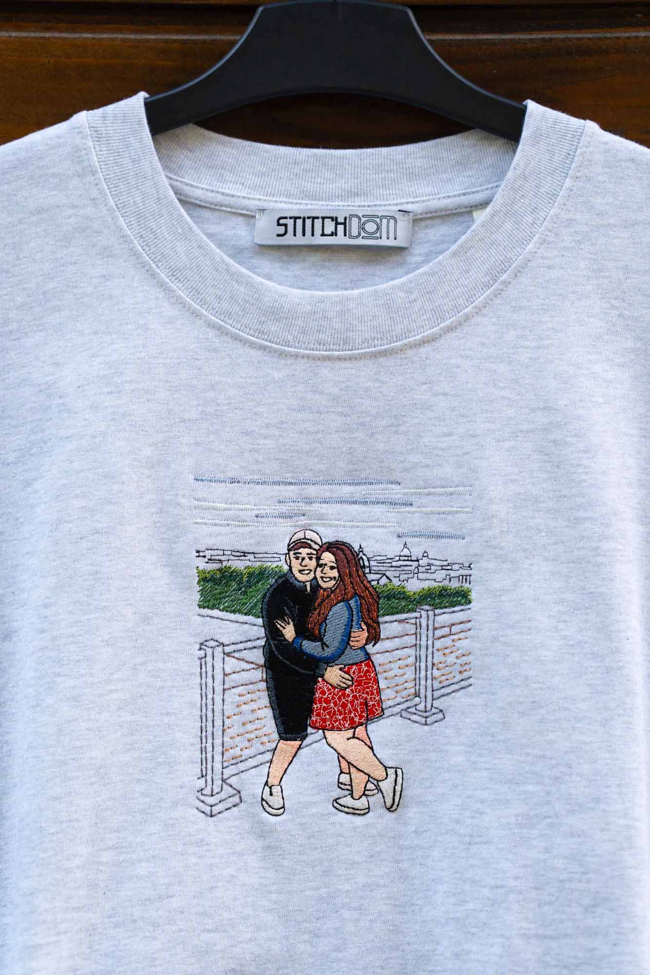 Tricou gri deschis din bumbac organic, pe umeraș negru, pe fundal din lemn închis, cu un model brodat personalizat reprezentând un cuplu care se îmbrățișează.
