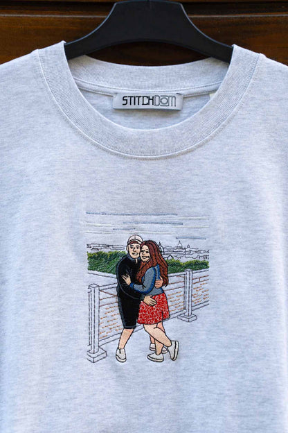 Tricou gri deschis din bumbac organic, pe umeraș negru, pe fundal din lemn închis, cu un model brodat personalizat reprezentând un cuplu care se îmbrățișează.