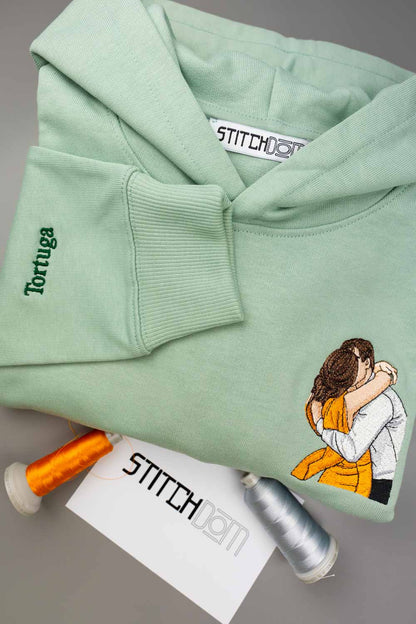 Hanorac verde mentă din bumbac organic, personalizat, cu textul „Tortuga” brodat pe mânecă și o poză brodată în stil desen cu un cuplu pe partea dreaptă jos a feței, alături de conuri de ață și un card cu textul „STITCHDOM”, pe un fundal gri neutru.
