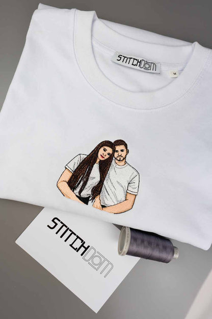 Tricou alb din bumbac organic, împăturit, cu o poză brodată personalizată în stil desen cu un cuplu pe piept, alături de o etichetă de brand „Stitchdom”, un card „Stitchdom” și un con de ață, pe un fundal gri.