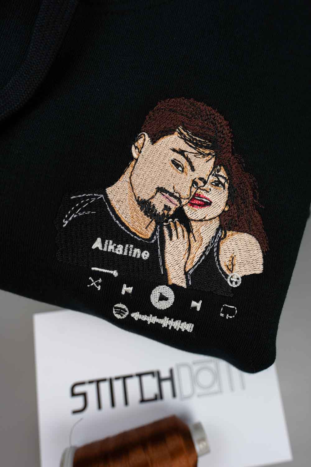 Hanorac negru din bumbac organic, cu o poză brodată de cuplu personalizată cu un cuplu, textul „Alkaline” și pictograme de player muzical, alături de un card alb cu brandul „StitchDOM” și un con de ață maro.