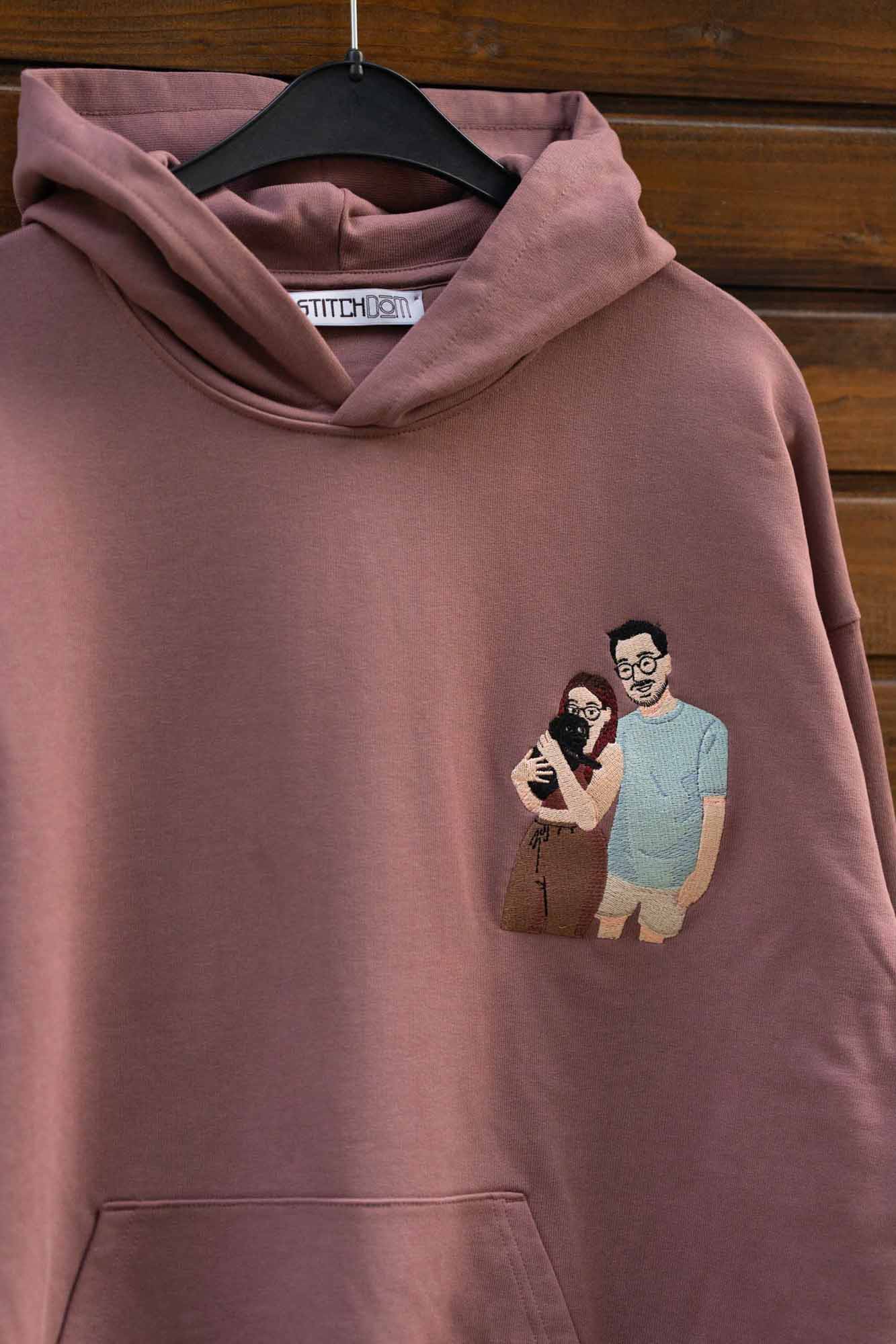 Hanorac maro din bumbac organic, pe umeraș negru, pe fundal din lemn, cu un model brodat personalizat pe partea stângă a pieptului reprezentând un cuplu care se îmbrățișează impreuna cu un catel.