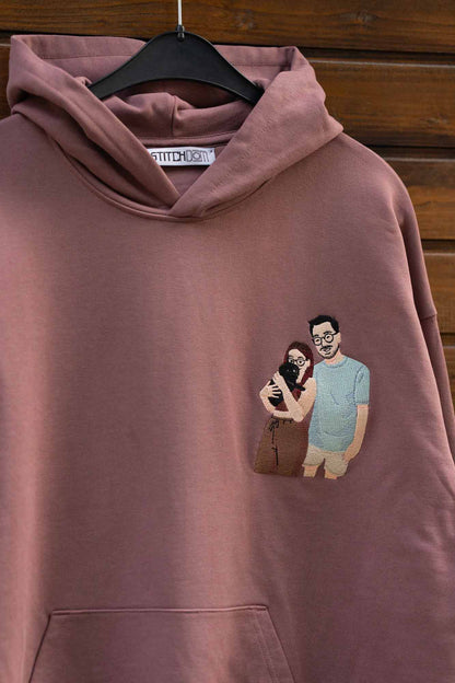 Hanorac maro din bumbac organic, pe umeraș negru, pe fundal din lemn, cu un model brodat personalizat pe partea stângă a pieptului reprezentând un cuplu care se îmbrățișează impreuna cu un catel.