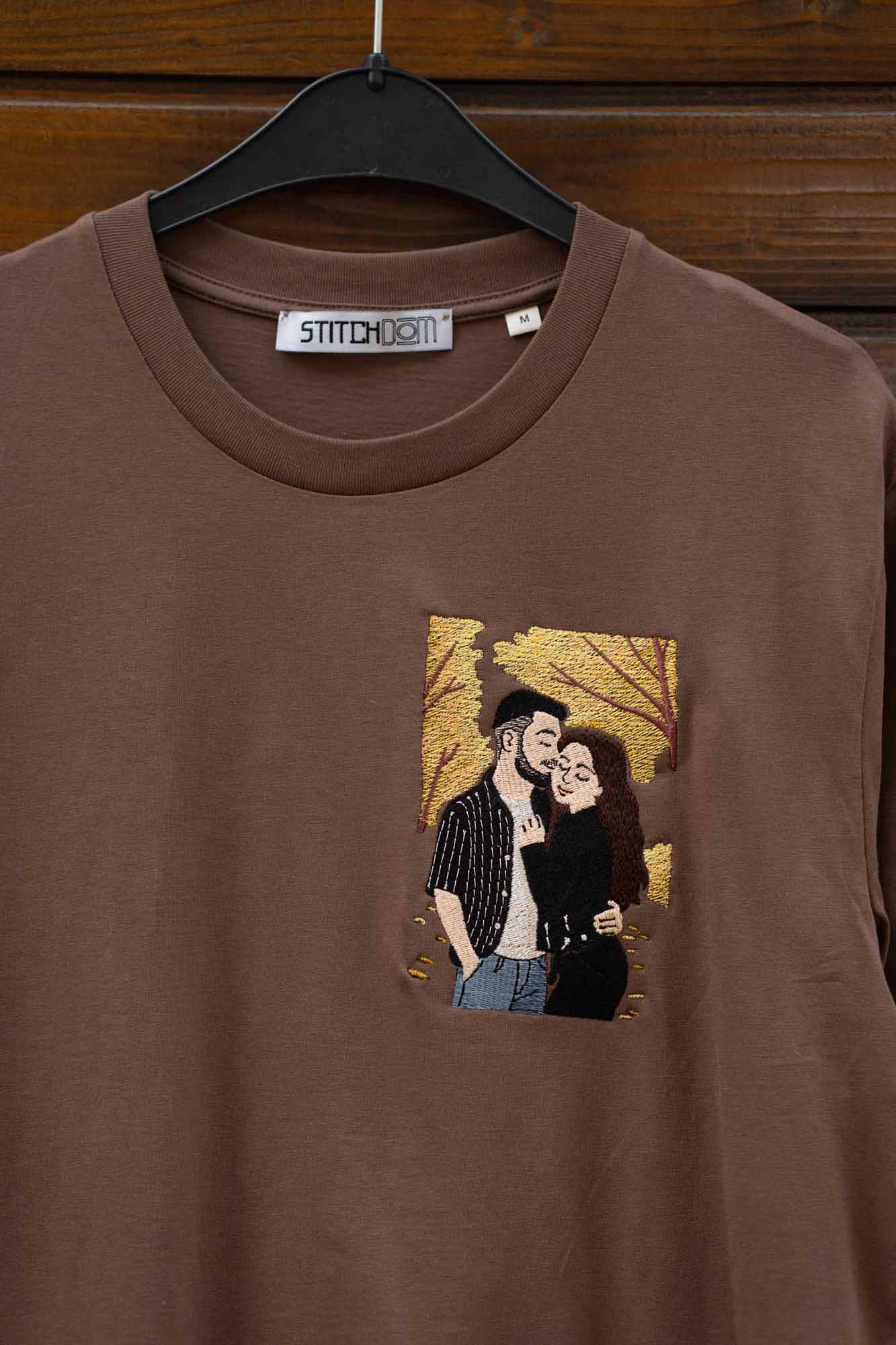 Tricou maro din bumbac organic, cu guler rotund, pe umeraș negru, pe fundal din lemn închis, cu un model brodat personalizat reprezentând un cuplu, pe un fundal cu model de frunze aurii.