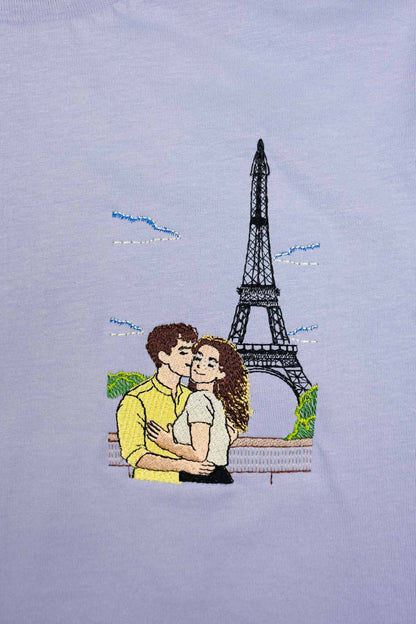 Tricou lavender din bumbac organic, cu un model brodat reprezentând un cuplu îmbrățișat în fața Turnului Eiffel, cu nori albaștri și detalii de vegetație verde.