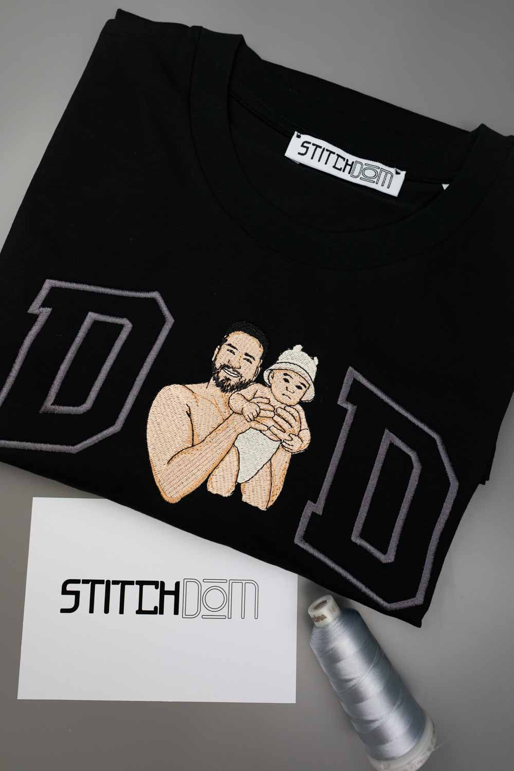 Tricou negru din bumbac organic, împăturit, cu o poză brodată personalizată cu un tată care își ține bebelușul în brațe, încadrat de două litere mari „D” conturate cu gri, cu eticheta „Stitchdom” vizibilă în interiorul gulerului, alături de un card „Stitchdom” și un con de ață, pe o suprafață gri.