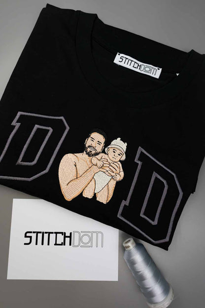 Tricou negru din bumbac organic, împăturit, cu o poză brodată personalizată cu un tată care își ține bebelușul în brațe, încadrat de două litere mari „D” conturate cu gri, cu eticheta „Stitchdom” vizibilă în interiorul gulerului, alături de un card „Stitchdom” și un con de ață, pe o suprafață gri.