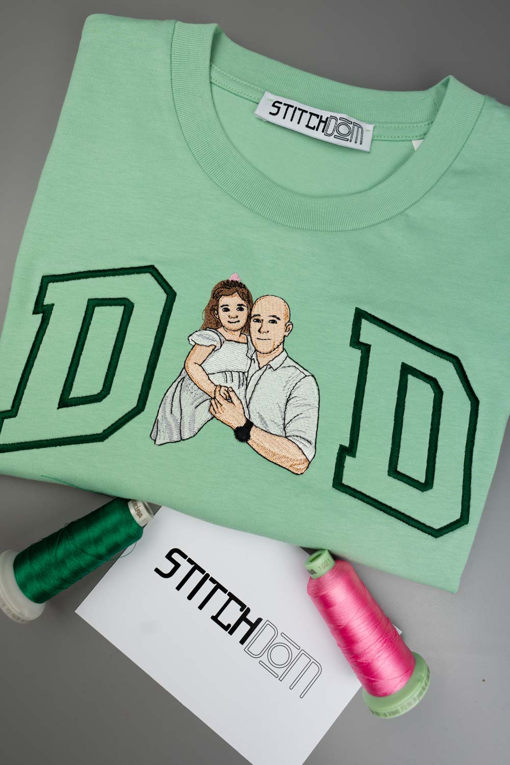 Tricou verde mentă din bumbac organic, cu o poză brodată personalizată în stil desen cu un cuplu, alături de două conuri de ață verde și roz și un card de brand „Stitchdom”, pe un fundal gri.