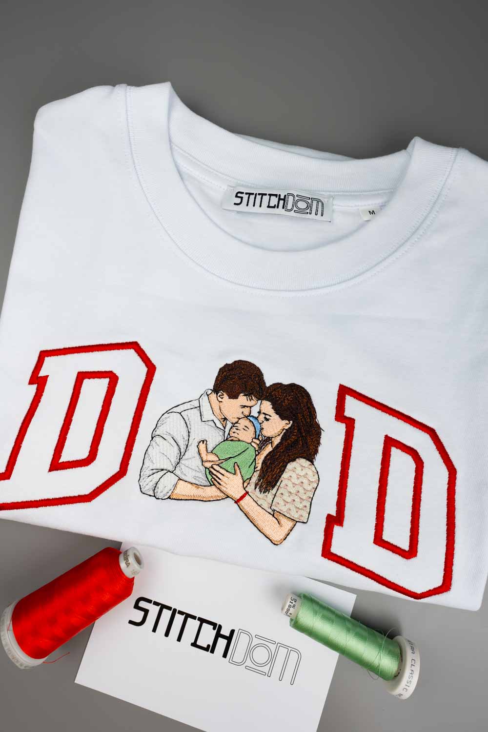 Tricou alb din bumbac organic, împăturit, cu literele mari „DAD” brodate cu contur roșu și o poză brodată cu un cuplu care ține un bebeluș, alături de conuri de ață și un card cu brandul „STITCHDOM”, pe un fundal gri.