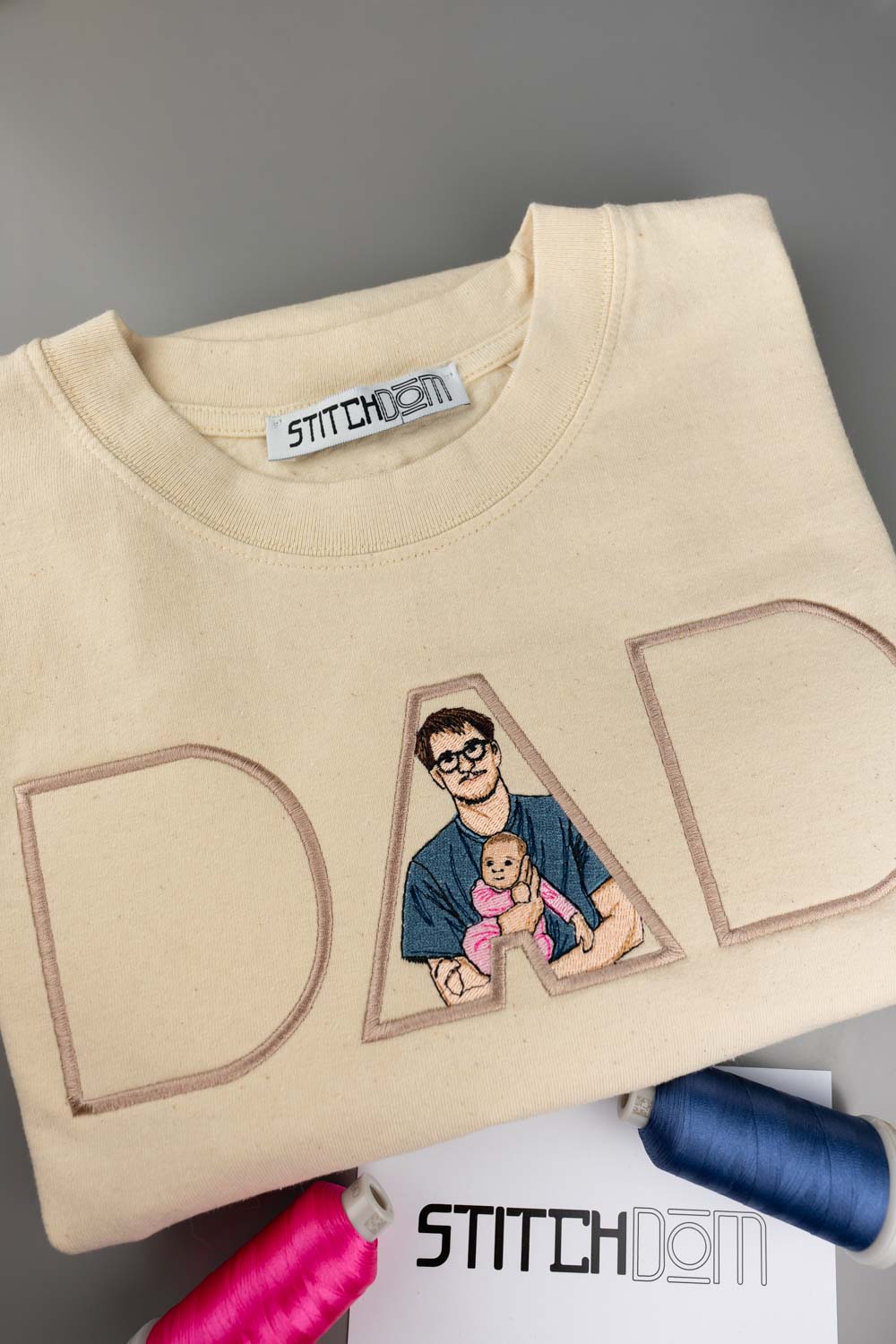 Tricou crem din bumbac organic, împăturit, cu un design brodat personalizat care formează textul „DAD” din litere mari conturate, litera „A” fiind umplută cu o ilustrație brodată reprezentând un tată care își ține bebelușul în brațe, pe o suprafață gri, alături de conuri de ață și o etichetă de brand „Stitchdom”.
