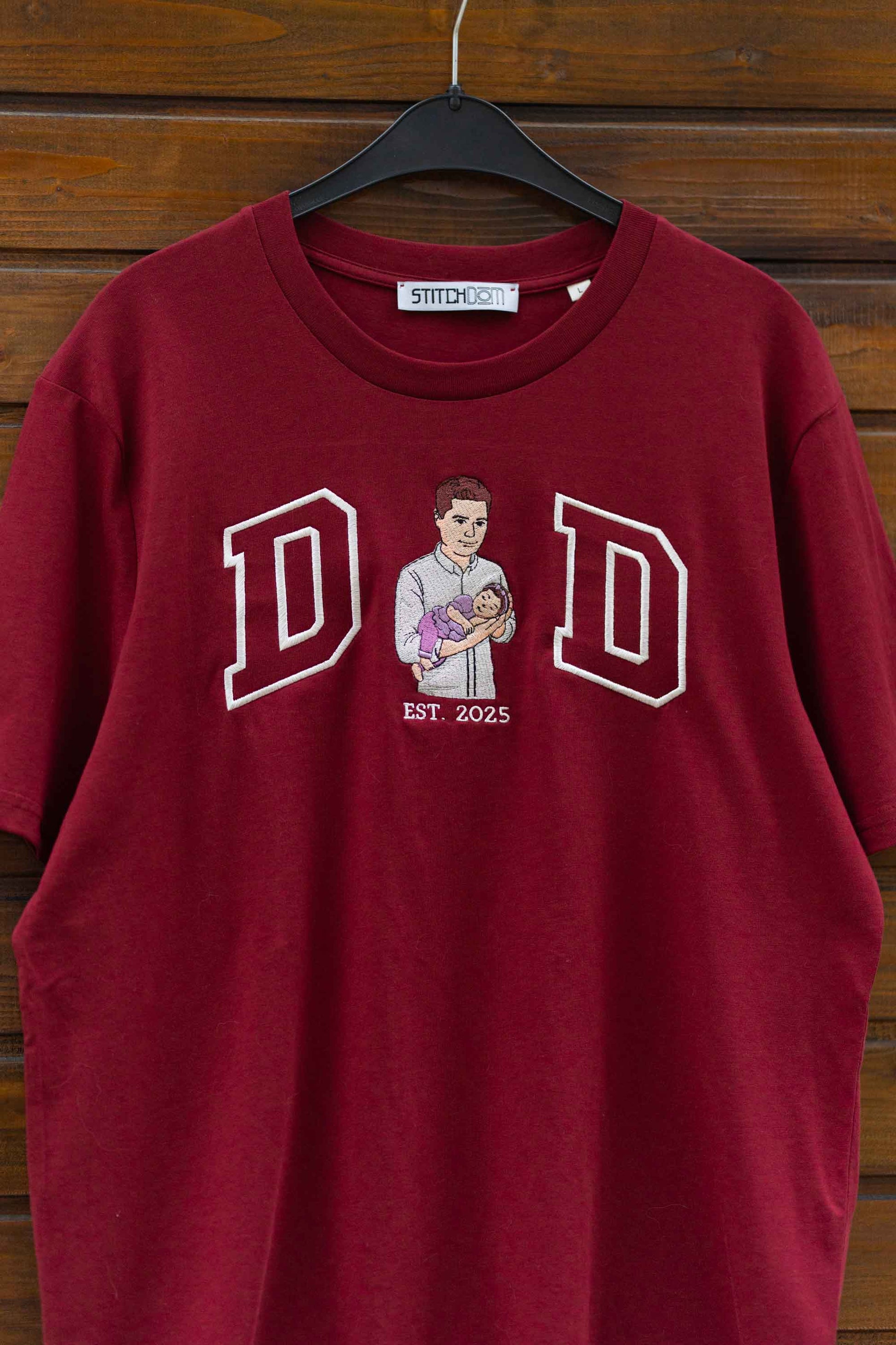 Tricou roșu închis din bumbac organic cu textul „DAD” și o poza brodata, pe un fundal din lemn.