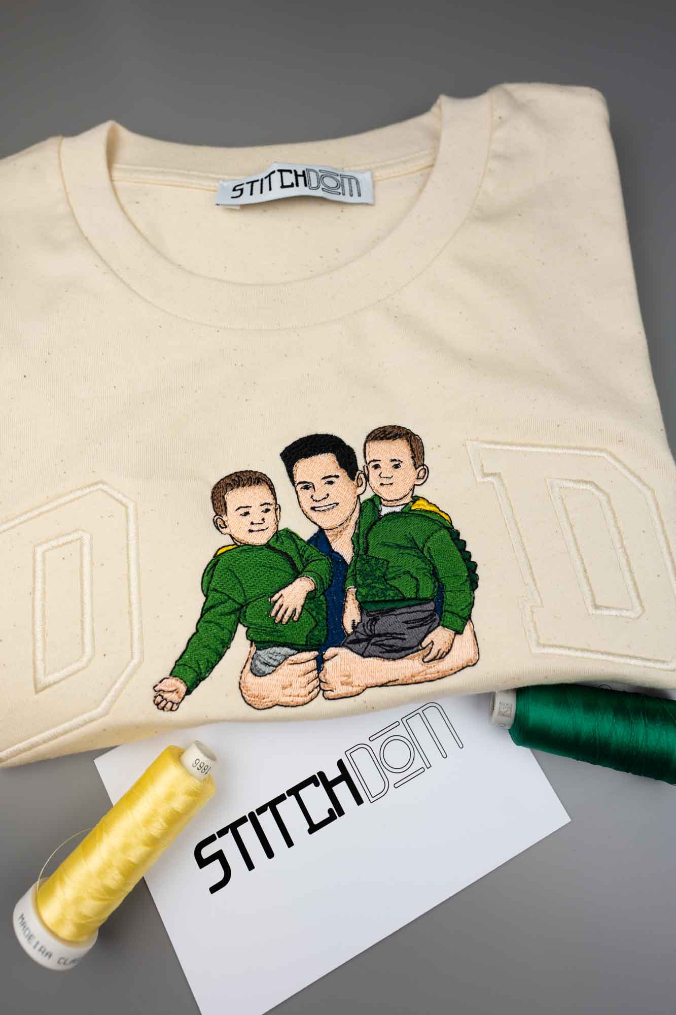Tricou crem din bumbac organic, împăturit, pe un fundal gri, cu o poză brodată personalizată cu trei bărbați așezați împreună, alături de conuri de ață și un card „Stitchdom”.
