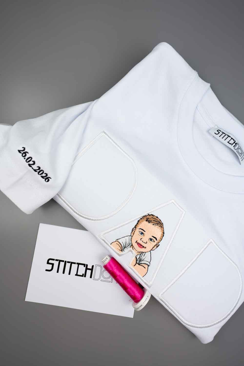Tricou alb din bumbac organic, împăturit, cu textul „DAD” și o poză brodată cu copilul, alături de un con de ață roz pe un card de brand, pe un fundal gri.