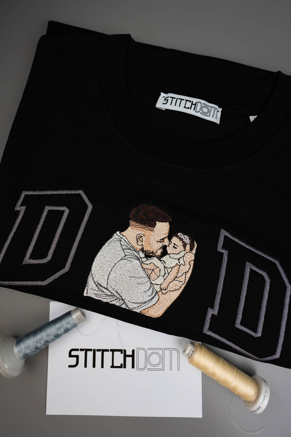 Tricou negru din bumbac organic, împăturit, cu o poză brodată personalizată cu un tată care își ține copilul în brațe, încadrat de două litere mari „D” conturate cu gri, alături de conuri de ață și o etichetă albă cu brandul „STITCHDOM”, pe un fundal gri.